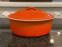Side view of the orange cast iron Dutch oven with lid on black countertop showing shape and color.