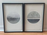 Two framed photographic prints with circular foggy forest imagery side-by-side against a white wall on the floor