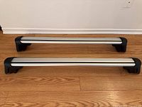 Full view of both silver metallic roof rack cross bars with black plastic end caps showing the length and general condition of the bars placed on wooden floor.