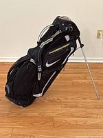 Side view of Nike golf bag standing on its metal legs on wooden floor, showing the side pockets and strap.