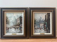 Pair of framed oil paintings on canvas signed Celestin, showing Paris street scenes with dark wooden frames and brass nameplates.
