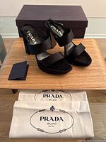 Photo showing the pair of Prada black satin heeled sandals with the original purple box in the background and two branded cloth dust bags in front.