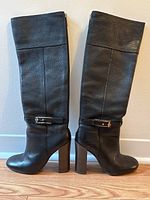 Side view of pair of tall black leather boots with block heels and gold buckle hardware.