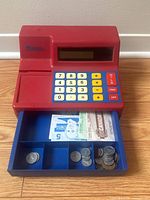 Front view of red and blue toy cash register with open cash drawer revealing assorted play money bills and coins.