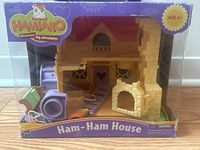 Front view of boxed Hamtaro Ham-Ham House playset showing beige plastic house structure with purple and green furniture pieces inside clear window packaging.