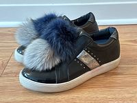Side angled view of black leather and vinyl sneakers with rabbit fur pom poms, metallic accents, and white rubber sole.