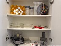 Kitchen cabinet shelf holding various kitchen items including Cuisinart food processor base, Pyrex casseroles, plastic containers, measuring tools, spice rack, and tin container.