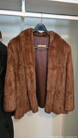 Front view of the brown waist length fur coat hanging on a black hanger showing open coat front and interior lining.