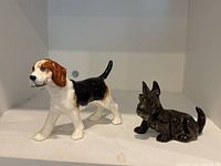 Two dog figurines displayed together on a white shelf: a tri-colored Beagle figurine and a black Scottie dog figurine.