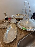 Entire assortment of silver-plated serving pieces displayed on a countertop, including platters, covered dish, trophy, candle holder, and red glass pot.