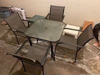 Top view of the square glass top metal frame outdoor table surrounded by four mesh fabric armchairs and two small plastic side tables placed on concrete floor.