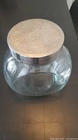 Top-down angled photo of the glass jar showing clear glass body and metal lid with embossed text.