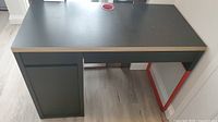 Full front view of the black IKEA MICKE desk showing the desk top, cabinet door on the left, and wood grain edges on the desk surface.
