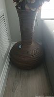Medium height brown rattan vase, coiled design, shown from a frontal angle on the floor near a window and wall.
