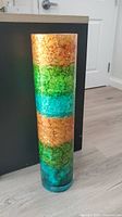 Tall cylindrical decorative glass vase shown standing upright against furniture on a wood floor, revealing color bands of amber, green, turquoise, and teal with crackled texture.