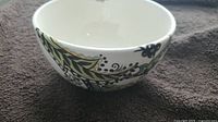 White ceramic bowl with intricate green and black floral pattern from side view on gray fabric.