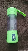 Front view of green Supkitdin portable blender showing the on/off button, clear blending container, and wrist strap attached to the lid.