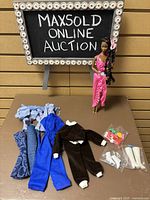 Shows Barbie doll standing in pink outfit with braided hair and pink ribbon, surrounded by four outfits and accessories on table.