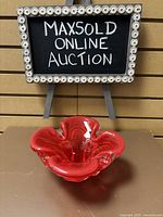 Front view of the red art glass dish, showing its wavy rim and vibrant red color with clear glass base accents, set against a wooden backdrop with a MaxSold sign.