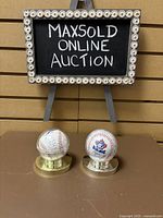 Photo showing both collectible baseballs in their clear plastic display cases with gold bases under a sign.