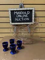 Copper wire chicken egg basket with four blue ceramic egg cups placed in front of a hanging chalkboard sign for MaxSold Online Auction.