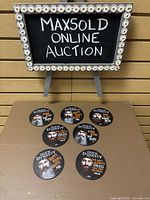 Set of seven round Duck Dynasty themed plastic coasters featuring character portraits and quotes, displayed under a sign reading 'MAXSOLD ONLINE AUCTION'