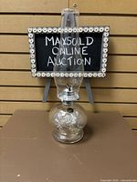 Front view of the clear glass oil lamp with hurricane shade on a shelf with a small sign behind.