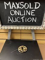 Brooch displayed pinned on a black surface under MaxSold Online Auction sign providing overall view of item size and style.