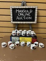 Full display of 23 ceramic NHL mini mugs showing various team logos arranged in two rows in front of a sign reading 'MAXSOLD ONLINE AUCTION'