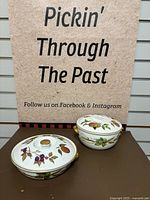 Two covered Royal Worcester porcelain casserole dishes with fruit and leaf decorations on lids and bodies, displayed on table with a backdrop.
