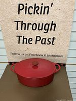 Front view of red oval Dutch oven with lid and side handles, placed against a background with text.