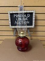 Full view of red glass oil lamp with clear hurricane chimney and brass burner in front of auction sign.