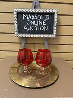 Two red glass snifters on a round brass charger plate with auction sign in background