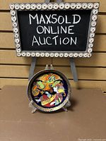 Full view of the Rosenthal Aladdin collectors plate on a display stand in front of MaxSold auction sign