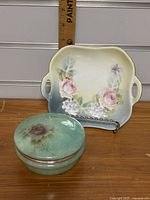 Photo of the green alabaster trinket box with lid showing a pink rose design, placed beside the German floral dish with handles, with a ruler for size reference.