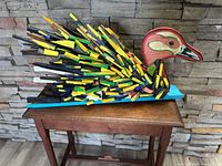 Full side view of the colorful wooden bird sculpture on a brown wooden table against a stone wall background.