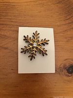 Gold-tone snowflake brooch with six branches and clear gemstone center on white display card.