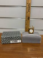 Wide shot of the Bulova mini alarm clock next to a ruler for size reference, placed on a grey stand, with the Bulova box beside it.