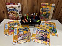 Photo showing 6 flattened Post Shreddies cereal boxes behind a display of 6 PeZ candy dispensers with colorful character heads, and 5 small toy cars positioned in front of the dispensers.