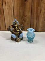 Resin Ganesh figure and blue hobnail vase placed side by side on a flat surface with wood panel background, showing full view of both items.