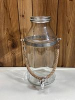 Full view of the tall glass beverage dispenser showing clear body, metal rim, rope handle, and spigot.
