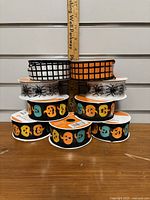 Front view of stacked nine rolls of Halloween ribbon with a ruler for scale showing 1.5 inch width of ribbons. Designs include checkered patterns, spider prints, and jack-o'-lantern faces.