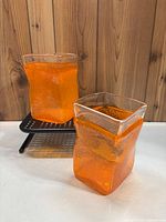Two heavy rectangular orange glass vases with clear irregular top rims and visible bubbles inside the glass, displayed on a white surface with wooden background