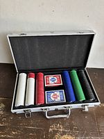 Open silver metal carrying case showing five stacks of plastic poker chips in different colors with two decks of Bicycle playing cards inside.