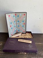 Scrabble game set displayed with foldable board standing inside box with letter tiles and tile holders inside box