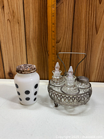 Front view of milk glass shaker with black dots and rusty lid next to metal cruet holder with four glass bottles.