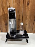 Silver SodaStream soda maker with black top panel and clear bottle beside it on black perforated stand.