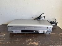 Front view of Sylvania 3-in-1 DVD, CD, VHS player showing power cord attached, placed on wooden surface with white background.