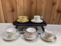 Photo showing five vintage teacups and saucers arranged in two rows on a black perforated tray and a wood surface.