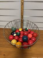 Photo of a silver wire basket with various colored vintage billiard balls inside, including multiple red balls and a few larger colored balls.